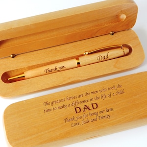 May include: A wooden pen and case set. The pen is engraved with "Dad" and "Thank you". The case is engraved with a message. The set is made of light-colored wood with gold accents.