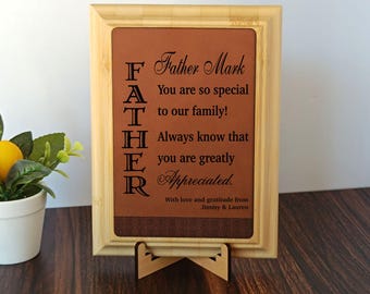 May include: A framed wooden plaque with a brown leather-like insert. The text reads "FATHER" vertically, with "Father Mark" above and a message of appreciation below. Includes a wooden stand.