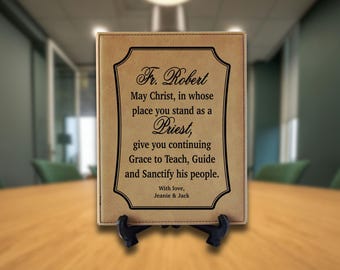 May include: A rectangular tan faux leather plaque with black stitched edges, displayed on a black plastic stand. The plaque features black engraved text: Fr. Robert, May Christ, in whose place you stand as a Priest, give you continuing Grace.