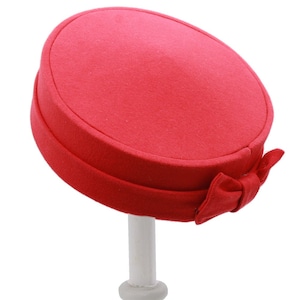 May include: A bright red pillbox hat with a bow on the side. The hat is made of a soft, felt-like material.