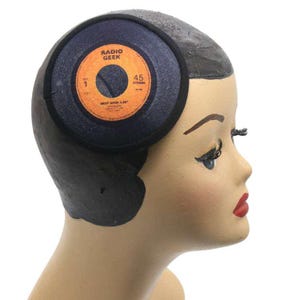 Könnte beinhalten: Ein Schaufensterpuppenkopf mit einer schwarzen Kappe mit einem kreisförmigen Design, das einer Schallplatte ähnelt. Die Schallplatte hat orangefarbene und schwarze Details mit dem Text "RADIO GEEK" und "45 STEREO". Die Schaufensterpuppe hat dunkles Haar, roten Lippenstift und lange Wimpern.