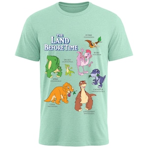 May include: A light green short-sleeve t-shirt featuring a graphic print of The Land Before Time characters including Spike, Ducky, Petrie, Ruby, Chomper, Cera, and Littlefoot with their names and species printed in blue and black text.