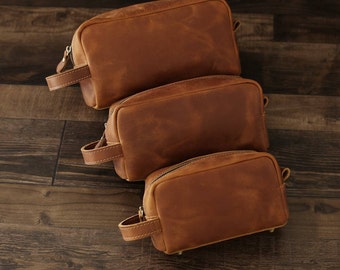 Peut inclure: Trois pochettes en cuir marron à fermeture éclair de différentes tailles empilées les unes sur les autres sur une surface en bois.