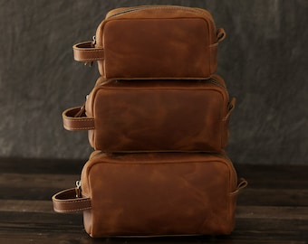 Peut inclure: Trois pochettes zippées en cuir marron empilées les unes sur les autres. Les pochettes sont en cuir vieilli et ont une poignée en cuir marron.