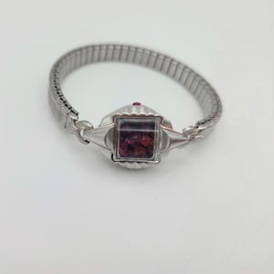 May include: A silver-toned stretch bracelet featuring a central square-shaped housing with a clear glass face. Inside the square compartment are small, loose, dark red and purple gemstone fragments. The band consists of flexible metal links.
