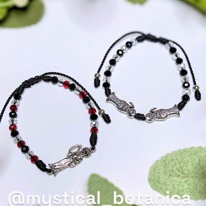 May include: Two black cord bracelets with red, clear, and black beads. Each bracelet features a silver-toned charm. The text "@mystical_botanica" is visible at the bottom of the image.