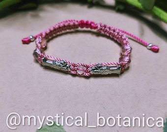 May include: Pink woven bracelet with silver-toned accents. The bracelet features a braided design with a central decorative element and adjustable cords. The text "@mystical_botanica" is visible at the bottom of the image.