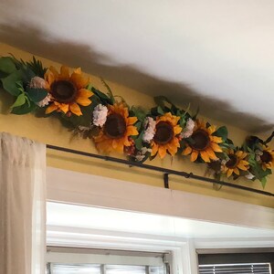 May include: A sunflower garland with green leaves and white flowers hangs from a curtain rod above a window with white blinds.