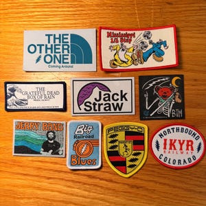 May include: A collection of embroidered patches with various designs and text. The patches feature imagery such as a wave, a bear skeleton, and text like "The Other One" and "Jack Straw". Colors include blue, red, yellow, and black.