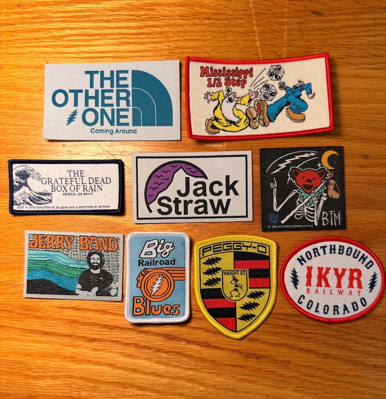 May include: A collection of embroidered patches with various designs and text. The patches feature imagery such as a wave, a bear skeleton, and text like "The Other One" and "Jack Straw". Colors include blue, red, yellow, and black.