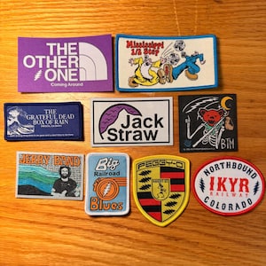 May include: A collection of embroidered patches with various designs and text. Patches include "The Other One," "Mississippi 1/2 Step," "The Grateful Dead Box of Rain," "Jack Straw," "Jerry Band," "Big Railroad Blues," "Peggy-O," and "Northbound IKYR Railway Colorado."