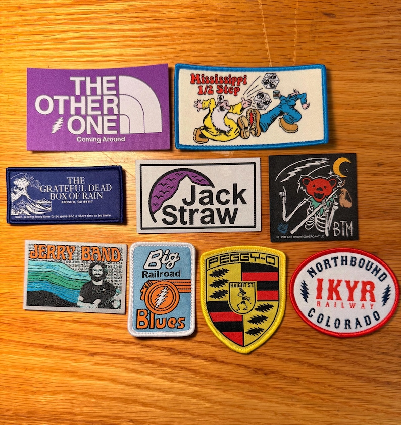 May include: A selection of embroidered patches with diverse designs and text. Patches feature "The Other One," "Mississippi 1/2 Step," "The Grateful Dead Box of Rain," "Jack Straw," "Jerry Band," "Big Railroad Blues," "Peggy-O," and "Northbound IKYR Railway Colorado."