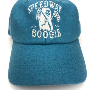 May include: A teal blue baseball cap with white embroidery. The embroidery features a skeleton holding a scythe and a checkered flag with the text "Speedway Boogie" underneath.