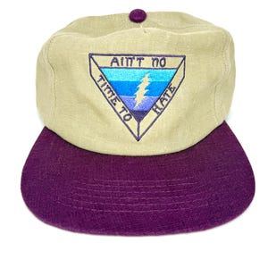 May include: A tan baseball cap with a purple brim and a purple button on top. The cap features an embroidered patch with the text "Ain't No Time To Hate" and a lightning bolt design in blue and purple.