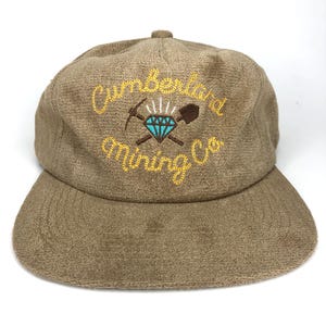 May include: A brown suede baseball cap with an embroidered logo that says "Cumberland Mining Co." in yellow lettering. The logo features a blue diamond with crossed pickaxes and a shovel.