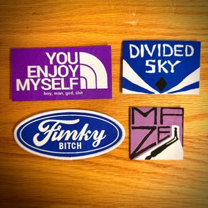 May include: Four embroidered patches on a wooden surface. The patches include text: "YOU ENJOY MYSELF", "DIVIDED SKY", "Funky Bitch", and "MAZE". The patches are in various colors, including purple, blue, and white.