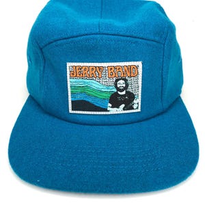 May include: A teal blue wool 5-panel cap with a patch featuring a photo of Jerry Garcia and the text "JERRY BAND" in orange and black letters.