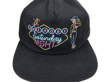 May include: Black baseball cap with a neon sign design that says "One More Saturday Night" and a cartoon cowboy in front of it.
