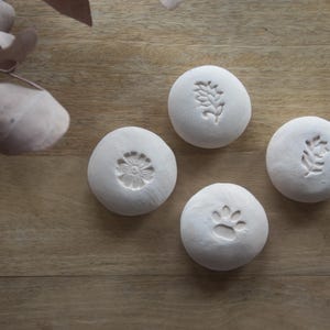 Könnte beinhalten: Fünf weiße Keramik-Untersetzer mit verschiedenen Designs. Die Designs beinhalten eine Blume, einen Pfotenabdruck und zwei verschiedene Blattmotive.