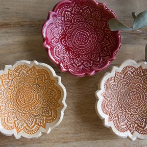 Könnte beinhalten: Drei dekorative Keramikschalen mit einem floralen Mandala-Design. Die Schalen sind in Rot-, Orange- und Brauntönen gehalten. Die Schalen haben einen gewellten Rand und stehen auf einer Holzoberfläche.