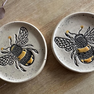 Könnte beinhalten: Zwei runde Keramikschalen mit gesprenkelter cremefarbener Oberfläche, die jeweils mit einem vertieften Hummelmotiv versehen sind. Die Bienen sind mit schwarzen Umrissen und gelben Akzenten an Hinterleib und Fühlern auf jeder Schale dargestellt.