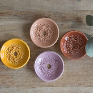 Könnte beinhalten: Vier Keramik-Seifenschalen mit aufwendigen floralen Designs. Die Schalen sind in den Farben Rosa, Orange, Gelb und Lila.