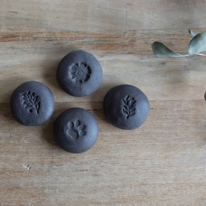 Könnte beinhalten: Vier runde schwarze Keramik-Untersetzer mit verschiedenen Designs. Die Designs beinhalten eine Blume, ein Blatt, einen Pfotenabdruck und einen Zweig.