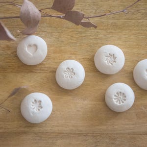 Könnte beinhalten: Sechs kleine, runde, cremefarbene Keramiksteine mit geprägten Designs, darunter ein Herz, ein Pfotenabdruck und florale Muster. Sie liegen auf einer Holzoberfläche mit getrockneten Blättern. Die Designs sind dezent erhaben.