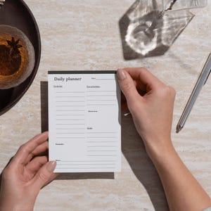 May include: A white daily planner with a black border, held by hands. The planner includes sections for to-do lists, top priorities, motivation, goals, reminders, and notes. The words "Daily planner" are printed at the top.