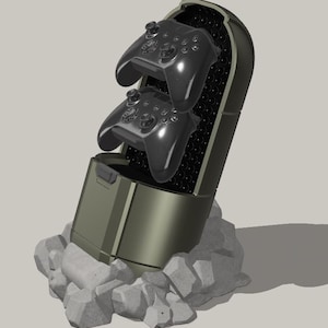 May include: An olive green gaming controller holder with two black controllers inside. The holder is set on a pile of grey rocks. The controllers are glossy and have multiple buttons and joysticks.