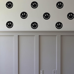 May include: Black smiley face wall decals are arranged on a white wall. The decals are evenly spaced and create a cheerful, playful design. The wall has a wainscoting detail at the bottom.