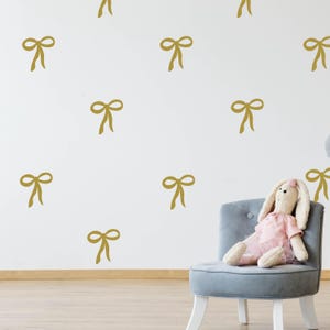May include: A white wall with a repeating pattern of gold bow decals. A gray chair with a stuffed bunny sitting on it.
