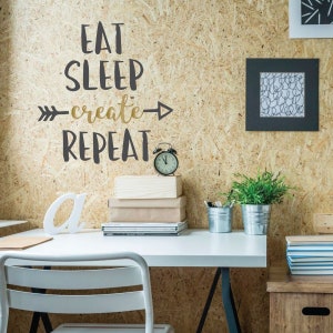 May include: A white desk with a black metal frame and a white chair. On the desk is a stack of books, a small alarm clock, two potted plants, and a white letter "a". The wall behind the desk is a light brown wood panel with a black and gold vinyl decal that reads "Eat Sleep Create Repeat" with an arrow pointing to the right.