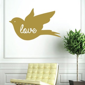 May include: Gold vinyl wall decal of a stylized bird with the word "love" written inside. The decal is on a white wall with a yellow chair and a green plant in a black pot.