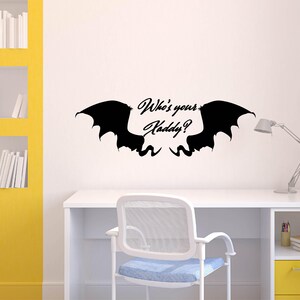 May include: A black wall decal featuring stylized bat wings with the text "Who's your Daddy?" in a cursive font. The decal is mounted on a white wall above a white desk with a chair and a desk lamp.