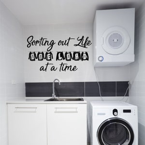 May include: A white laundry room with a washing machine, dryer, and a sink. The wall has a black vinyl decal that reads "Sorting out life one load at a time".
