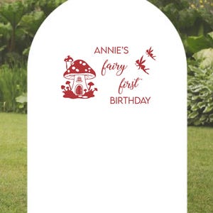 May include: White arch with red text and a mushroom house illustration. The text reads "Annie's fairy first birthday".