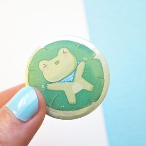 Pode incluir: Um botão verde e branco com um sapo de desenho animado usando uma bandana quadriculada azul e branca, deitado em um lírio d'água verde.