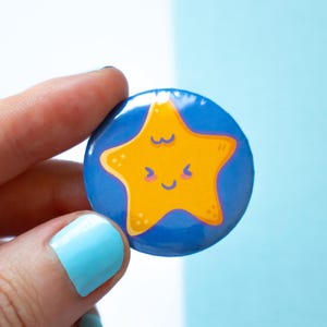 May include: A round, blue button with a yellow cartoon star design. The star has a smiling face and is surrounded by a blue background. The button is held by a hand with light blue nail polish.