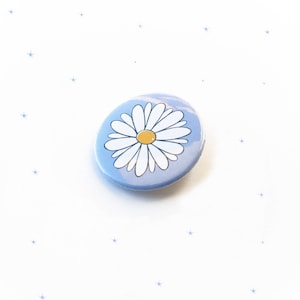 Pode incluir: Um botão azul e branco com uma margarida branca com um centro amarelo. O botão está sobre um fundo branco com pequenas estrelas azuis.