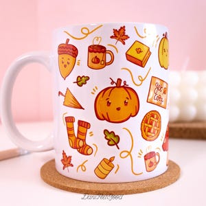 Pode incluir: Caneca de cerâmica branca com um design temático de outono. A caneca apresenta ilustrações de abóboras, folhas, bolotas, uma torta, um livro e uma xícara de café. O texto "Get Cozy" também está incluído na caneca.