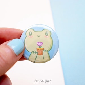 Pode incluir: Um botão redondo com um desenho de um sapo de desenho animado segurando uma flor num vaso. O sapo é verde claro com bochechas rosadas e uma expressão sorridente. O botão tem um fundo azul claro e o texto "DaniFeelsGood" na parte inferior.