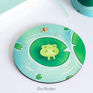 Può includere: Un sottobicchiere rotondo in legno con un design ad acquerello blu e verde. Una rana verde sta dormendo su una ninfea al centro del sottobicchiere. Il testo "Dani Feels Good" è stampato nell'angolo in basso a destra.