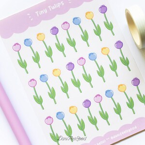 Pode incluir: Uma folha de autocolantes com filas de pequenas tulipas ilustradas em roxo, azul e amarelo com caules e folhas verdes. A folha tem uma borda recortada em lavanda no topo e na base, com o texto Tiny Tulips e DaniFeelsGood impresso.