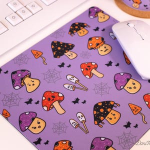 Pode incluir: Tapete de rato roxo com um padrão repetido de cogumelos de desenho animado com chapéus e caules de diferentes cores. Os cogumelos têm olhos e sorriem. Também há pequenos morcegos pretos e teias de aranha no tapete.