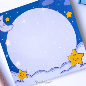 Puede incluir: Un bloc de notas cuadrado con un tema de cielo nocturno. El bloc de notas presenta un gran círculo blanco en el centro, rodeado por un borde azul con una luna creciente, estrellas y nubes. El bloc de notas tiene el texto "DaniFeelsGood".