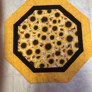 May include: An octagonal placemat featuring a sunflower pattern. The center of the placemat is filled with sunflowers, surrounded by a black border and a yellow patterned edge. The overall design is bright and cheerful.