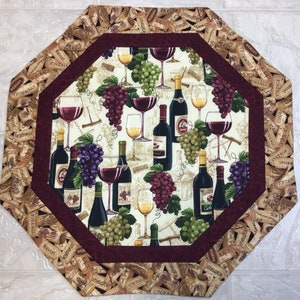 May include: Octagonal table runner with a burgundy border and a cream background. The runner features a repeating pattern of wine bottles, grapes, and wine glasses. The border is made of cork pieces with the words "Vintage", "Bordeaux", and "Merlot" printed on them.