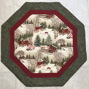 May include: Octagonal table runner with a red and green border. The center features a winter scene with a white background, red and green trees, and horses pulling a wagon.