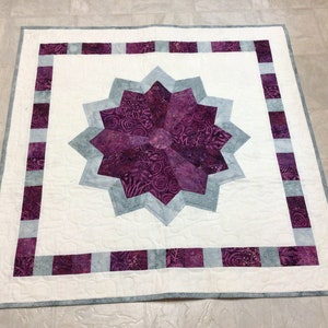 May include: A purple and gray quilt with a starburst design in the center. The quilt is surrounded by a border of purple and gray squares.